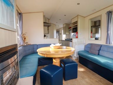 A dining area with a round wooden table surrounded by blue cushioned benches and stools next to a kitchen in Little Trevorrick Caravan in St Issey