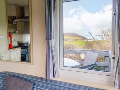A living area with a striped blue sofa and window showing an outdoor chair and table and countryside view at Little Trevorrick Caravan in St Issey