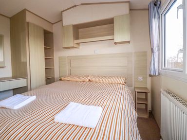 A bedroom with a double bed with striped bedding two towels on bed wooden cabinets and a window at Little Trevorrick Caravan in St Issey