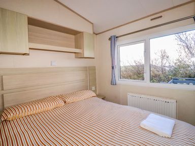 A bedroom with a double bed striped bedspread a window with curtains and wooden shelves at Little Trevorrick Caravan in St Issey