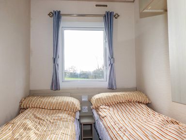 A small bedroom with two single beds striped bedding a window with curtains and a small bedside table at Little Trevorrick Caravan in St Issey