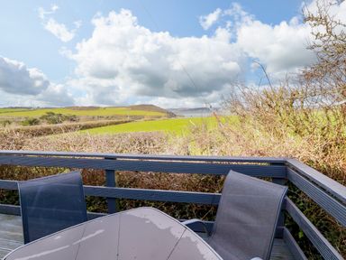 A balcony with a glass table and two chairs overlooking green fields and hills at Little Trevorrick Caravan in St Issey