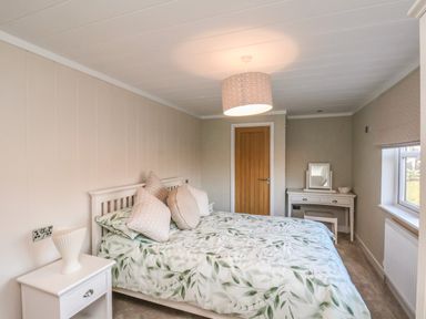 A bedroom with a bed with leafy patterned bedding a bedside table a dressing table a wooden door and a window at Cherry Blossom Lodge in Whitby