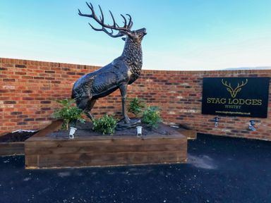 A statue of a stag on a raised wooden platform with plants in front of a brick wall and a sign that reads Stag Lodges Whitby