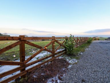 A wooden fence with string lights along a gravel path and grass fields at Cherry Blossom Lodge in Whitby