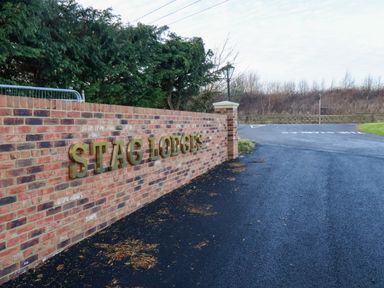 A brick wall with stag lodges sign beside a paved road and trees in the background at Cherry Blossom Lodge in Whitby