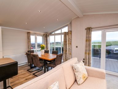 A dining area with wooden table and six chairs and a beige sofa with cushions near large windows and sliding doors at Holly Lodge in Whitby