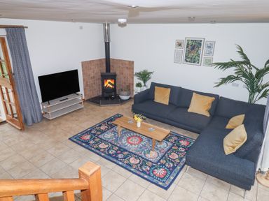 A living room with a dark gray sectional sofa and mustard pillows a wooden coffee table on a blue patterned rug a wood burning stove and a flat screen tv at The Old Coach House Little Dunley Cottages near Bovey Tracey