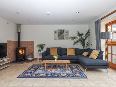 A living room with a dark sectional sofa and a coffee table on a patterned rug near a wood burning stove at The Old Coach House Little Dunley Cottages near Bovey Tracey