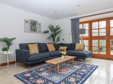 A living room with a blue sectional sofa with yellow cushions a wooden coffee table on a colorful patterned rug plants and framed pictures on the wall at The Old Coach House Little Dunley Cottages near Bovey Tracey