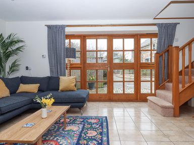 A living room with a blue sectional sofa yellow pillows a wooden coffee table with flowers a patterned rug near wooden framed glass doors and carpeted stairs at The Old Coach House Little Dunley Cottages near Bovey Tracey