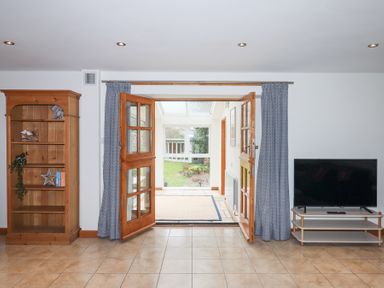 A room with wooden doors opening to a garden with a wooden bookshelf on the left and a television on the right at The Old Coach House in Little Dunley Cottages near Bovey Tracey