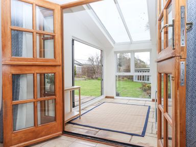 A room with wooden framed glass doors opening to a stone floor area with a rug and a small wooden table and large windows looking out to a garden at The Old Coach House Little Dunley Cottages near Bovey Tracey