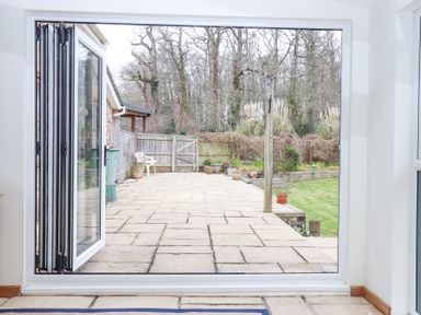 A patio with open glass doors leading to a garden with a lawn and trees at The Old Coach House in Little Dunley Cottages near Bovey Tracey
