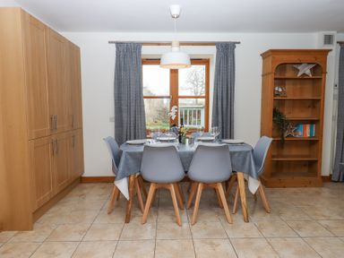 A dining area with a table set for six, grey chairs, wooden cabinets, and a wooden bookshelf at The Old Coach House Little Dunley Cottages near Bovey Tracey