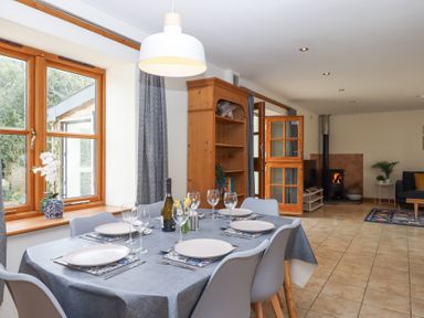 A dining area with a set table next to a window and an open door leading to a living room with a wood stove at The Old Coach House in Little Dunley Cottages near Bovey Tracey