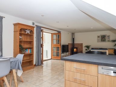 An open-plan kitchen and living area with wooden shelves dining table chairs and a wood stove at The Old Coach House Little Dunley Cottages near Bovey Tracey