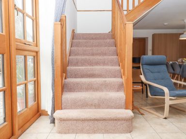 A carpeted staircase with wooden handrails next to a blue armchair and dining area at The Old Coach House Little Dunley Cottages near Bovey Tracey