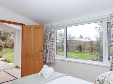 A bedroom with a bed, rolled towels, wooden door open to a garden, and large windows with floral curtains at The Old Coach House in Little Dunley Cottages near Bovey Tracey