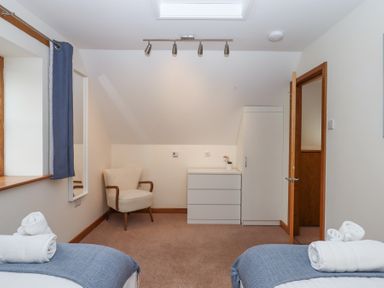 A bedroom with two beds, a beige armchair, a white chest of drawers, a white wardrobe, and a window with blue curtains at The Old Coach House Little Dunley Cottages near Bovey Tracey