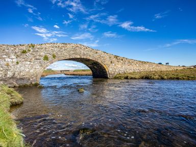 A stone arch bridge over a river with grassy banks under a blue sky at School House in Aberffraw