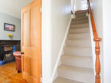 Carpeted staircase with wooden handrail next to an open wooden door showing part of a room with a fireplace leather chair and a vase with flowers at School House in Aberffraw