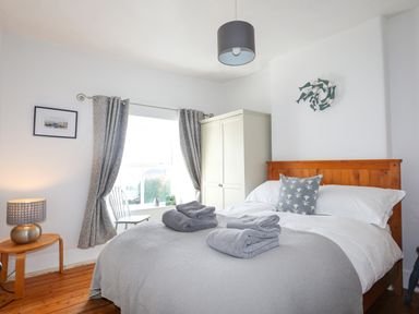 A bedroom with a wooden bed, gray bedding and towels, a white wardrobe, window with curtains, and a lamp on a wooden side table at School House in Aberffraw