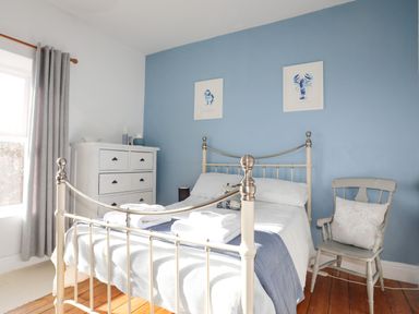 A bedroom with a metal bed frame white chest of drawers wooden chair and blue accent wall with paintings at School House in Aberffraw