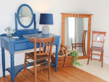 A room with a blue vanity table with an oval mirror and a wooden chair next to a large wooden-framed mirror and another wooden chair at School House in Aberffraw