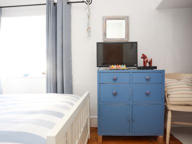 A bedroom with a white bed frame with striped bedding a blue dresser with a television and decorative items a chair with pillows and a window with blue curtains at School House in Aberffraw