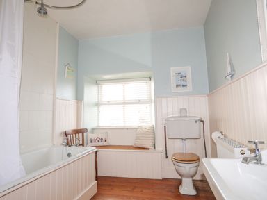 A bathroom with a bathtub a wooden chair a window seat with cushions a toilet and a sink at School House in Aberffraw