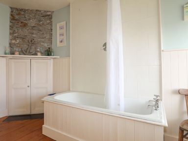 A bathroom with a white bathtub with shower curtain and silver taps a wooden chair and built in white cupboards with decorative bottles and an anchor at School House in Aberffraw
