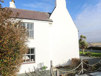 An exterior view of a white house with two windows a small outdoor seating area and plants at School House in Aberffraw