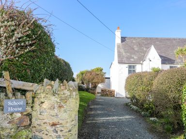 A gravel driveway leading to a white house with hedges on both sides and a stone wall with a School House sign at Aberffraw