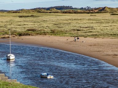 A river with boats near a sandy shore with two people walking dogs on the beach at School House in Aberffraw