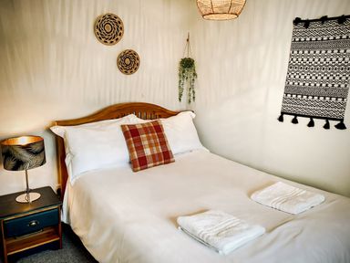 A bedroom with a wooden bed white bedding a checked pillow two folded towels a bedside table with a lamp and wall decorations at Hawthorn Crackington near Crackington Haven