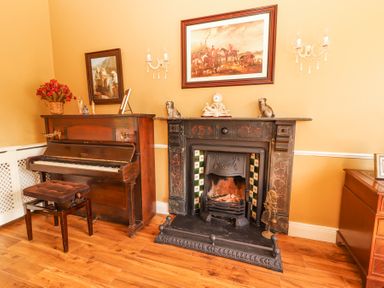 A room with a wooden piano and stool next to a black ornate fireplace with framed paintings on the wall at Dromclough Farmhouse in Dromclough near Listowel County Kerry