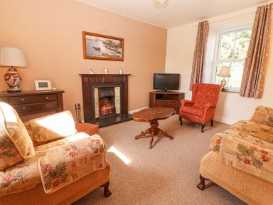 A living room with armchairs a wooden coffee table a fireplace a television on a wooden stand and a sideboard with a lamp and picture at Dromclough Farmhouse in Dromclough near Listowel County Kerry