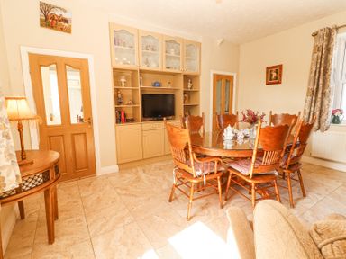 A dining room with a wooden table and six chairs a display cabinet with a television and decorative items a wooden door a small side table with a lamp and a window with curtains at Dromclough Farmhouse in Dromclough near Listowel County Kerry