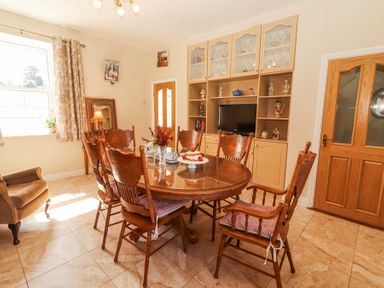 A dining room with a wooden table and six chairs a built-in cabinet with a television and ceramics an armchair and a window with curtains at Dromclough Farmhouse in Dromclough near Listowel County Kerry