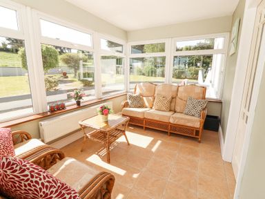 A sunroom with wicker furniture and cushions a small table in the center and large windows overlooking a garden at Dromclough Farmhouse in Dromclough near Listowel County Kerry