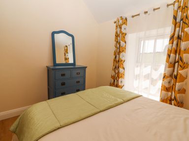 A bedroom with a bed, green blanket, blue dresser with mirror, and window with floral curtains at Dromclough Farmhouse in Dromclough near Listowel County Kerry
