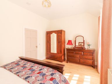 A bedroom with a wooden bed frame a dresser with a mirror and a red lamp a wooden wardrobe and pink curtains at Dromclough Farmhouse in Dromclough near Listowel County Kerry