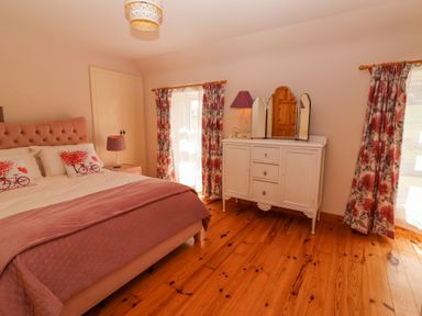 A bedroom with a bed with pillows and covers a nightstand with a lamp a white dresser with a three-panel mirror floral curtains and wooden floor at Dromclough Farmhouse in Dromclough near Listowel County Kerry
