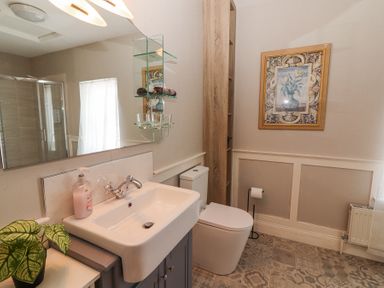 A bathroom with a white sink cabinet a toilet a framed painting a small plant and a glass shelf with towels and glasses at Dromclough Farmhouse in Dromclough near Listowel County Kerry