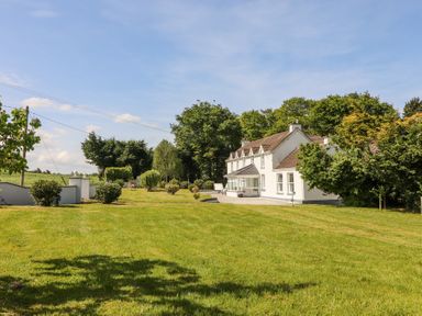 A white farmhouse with a tiled roof and a large lawn with bushes and trees at Dromclough Farmhouse in Dromclough near Listowel County Kerry