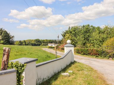 A rural road with a white wall and greenery alongside leading to a distant white house surrounded by trees at Dromclough Farmhouse Dromclough near Listowel County Kerry