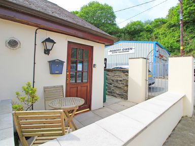 An outdoor patio area with wooden chairs and table by a brown door next to a white wall and gate at Fisherman Cove in Porthmadog