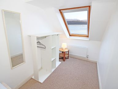 A small room with a white wardrobe, a round wooden side table with a lamp, a tall mirror on the wall, and a slanted window showing water and hills at Fisherman Cove in Porthmadog