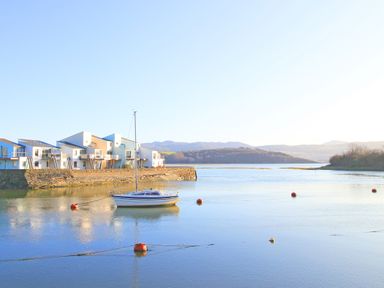 A sailboat moored by a stone pier with modern white buildings and hills in the background at Fisherman Cove in Porthmadog